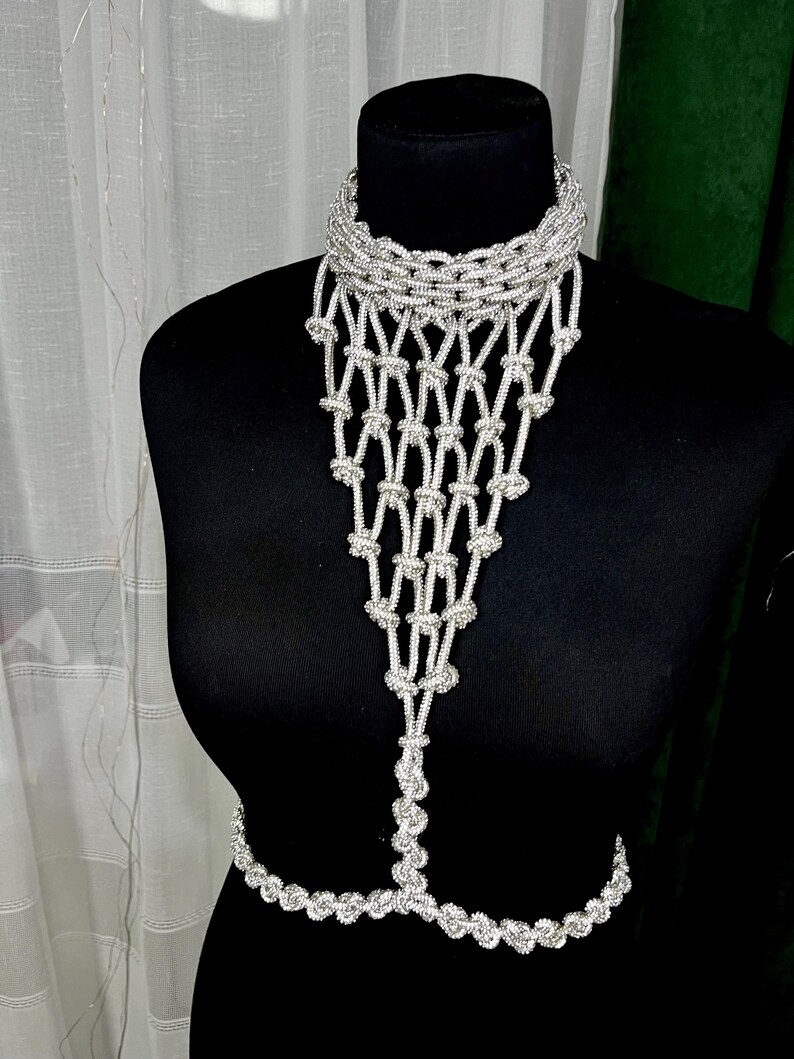 Puede incluir: Una joya corporal de cuentas plateadas que presenta un collar tipo gargantilla de cuello alto conectado a un babero en cascada de red anudada que se extiende hacia abajo hasta una cadena de cintura horizontal, hecha de peque&ntilde;as cuentas met&aacute;licas.
