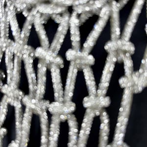 Puede incluir: Una tela de malla plateada con un patr&oacute;n de nudos repetitivo. El material tiene una textura superficial altamente reflectante y brillante que crea una estructura en forma de rejilla de bucles y nudos interconectados en toda el &aacute;rea visible.