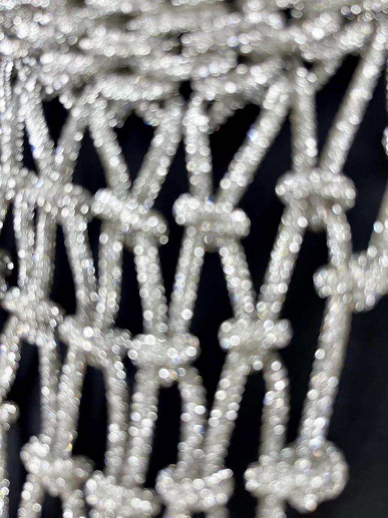 Puede incluir: Una tela de malla plateada con un patr&oacute;n de nudos repetitivo. El material tiene una textura superficial altamente reflectante y brillante que crea una estructura en forma de rejilla de bucles y nudos interconectados en toda el &aacute;rea visible.
