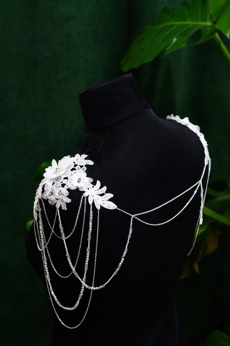 May include: A white lace shoulder piece with delicate floral designs and silver chains. The piece is displayed on a black mannequin against a green background.