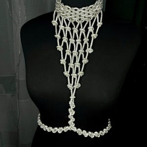 Puede incluir: Una joya corporal de tono plateado que presenta un collar tipo gargantilla conectado a un peto de macram&eacute; anudado en cascada, que se estrecha hacia una hebra vertical que se une a una cadena de cintura horizontal de eslabones trenzados.