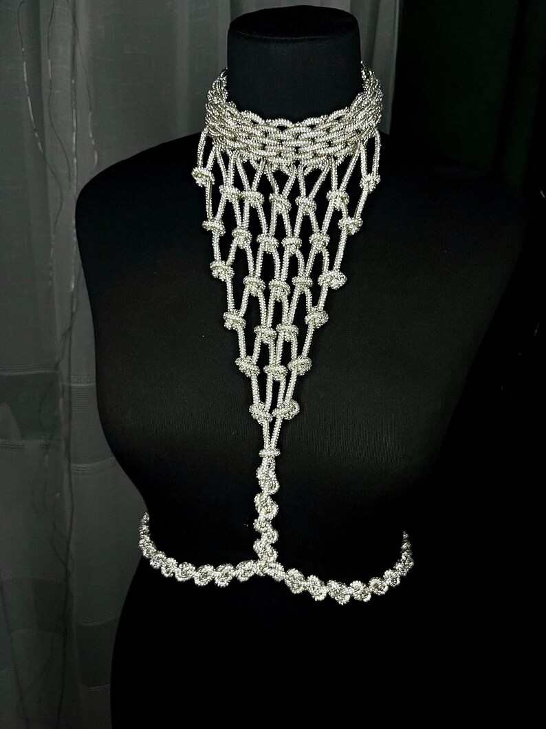 Puede incluir: Una joya corporal de tono plateado que presenta un collar tipo gargantilla conectado a un peto de macram&eacute; anudado en cascada, que se estrecha hacia una hebra vertical que se une a una cadena de cintura horizontal de eslabones trenzados.
