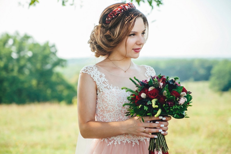May include: A bride in a pink and white lace dress holds a bouquet of red and green flowers. She is wearing a red and gold crown.