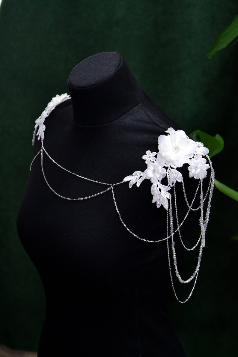 May include: White lace shoulder piece with delicate floral embellishments and a silver chain. The piece is displayed on a black mannequin.