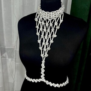 Puede incluir: Una joya corporal de tono plateado que presenta un collar tipo gargantilla conectado a un peto de red anudado en cascada que se extiende hacia abajo hasta una cadena que rodea la cintura. El dise&ntilde;o consta de eslabones y nudos plateados.
