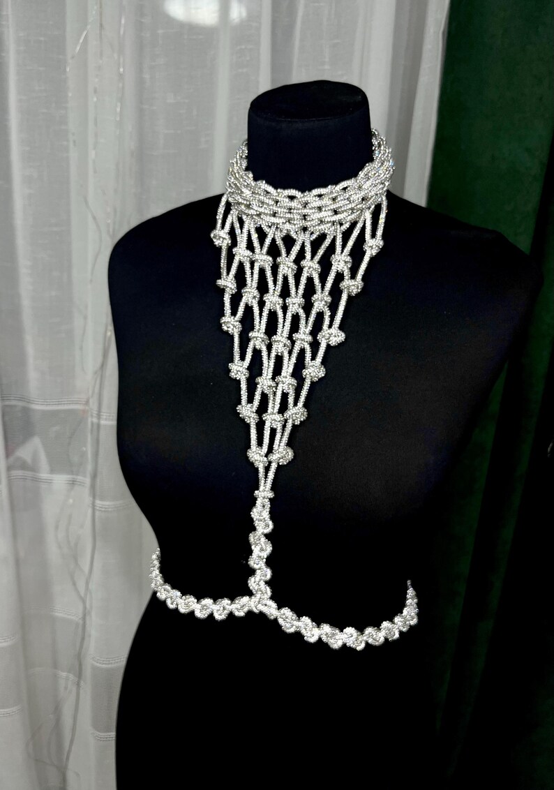 Puede incluir: Una joya corporal de tono plateado que presenta un collar tipo gargantilla conectado a un peto de red anudado en cascada que se extiende hacia abajo hasta una cadena que rodea la cintura. El dise&ntilde;o consta de eslabones y nudos plateados.
