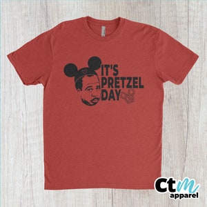 K&ouml;nnte beinhalten: Rotes T-Shirt mit einem schwarzen Aufdruck eines Mannes mit Mickey-Mouse-Ohren und dem Text "It's Pretzel Day" mit einem Brezel.