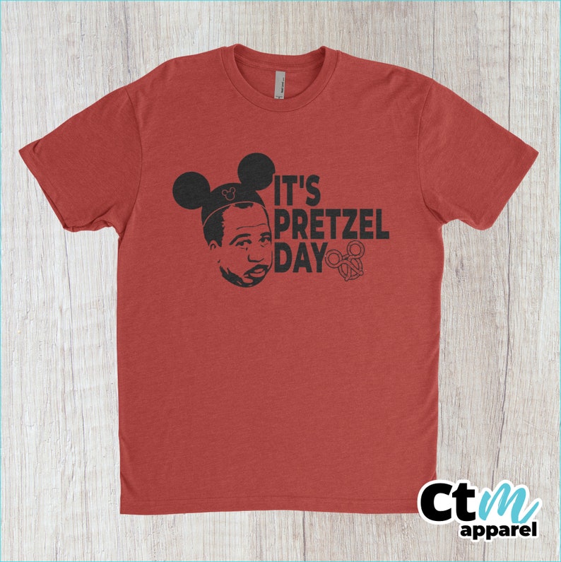 K&ouml;nnte beinhalten: Rotes T-Shirt mit einem schwarzen Aufdruck eines Mannes mit Mickey-Mouse-Ohren und dem Text "It's Pretzel Day" mit einem Brezel.