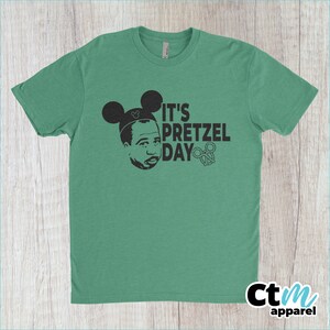 K&ouml;nnte beinhalten: Ein gr&uuml;nes T-Shirt mit einer schwarz-wei&szlig;en Grafik einer Person mit Mickey-Maus-Ohren und dem Text "It's Pretzel Day".