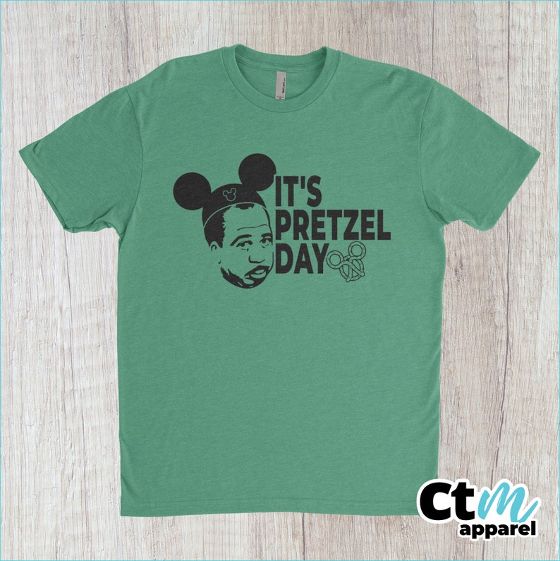 K&ouml;nnte beinhalten: Ein gr&uuml;nes T-Shirt mit einer schwarz-wei&szlig;en Grafik einer Person mit Mickey-Maus-Ohren und dem Text "It's Pretzel Day".