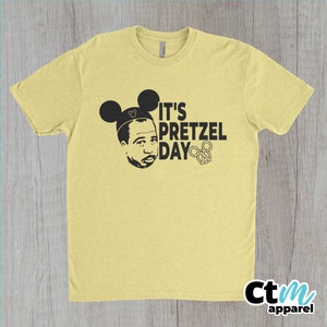 K&ouml;nnte beinhalten: Ein gelbes T-Shirt mit einer schwarzen Grafik einer Person, die Mickey-Maus-Ohren tr&auml;gt und den Text "It's Pretzel Day" mit einem Brezel.