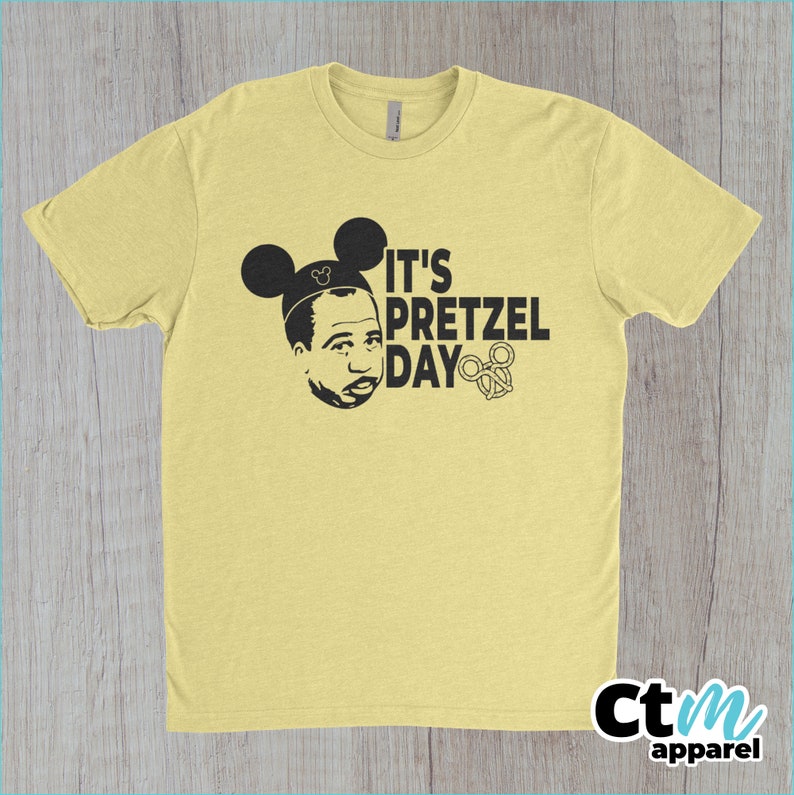 K&ouml;nnte beinhalten: Ein gelbes T-Shirt mit einer schwarzen Grafik einer Person, die Mickey-Maus-Ohren tr&auml;gt und den Text "It's Pretzel Day" mit einem Brezel.
