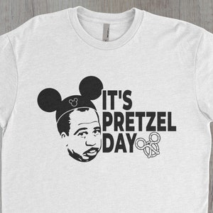May include: White t-shirt with a black and white graphic of a person wearing Mickey Mouse ears and the text "It's Pretzel Day".