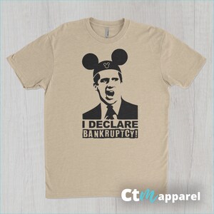 May include: A tan t-shirt with a black and white image of a man wearing Mickey Mouse ears. The man is wearing a suit and tie and has a surprised expression on his face. The text "I DECLARE BANKRUPTCY!" is printed below the image.