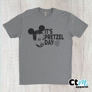 K&ouml;nnte beinhalten: Ein graues T-Shirt mit einer schwarzen Grafik einer Person, die Mickey-Maus-Ohren tr&auml;gt und den Text "It's Pretzel Day" mit einem Brezel-Symbol.