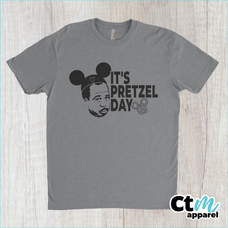 K&ouml;nnte beinhalten: Ein graues T-Shirt mit einer schwarzen Grafik einer Person, die Mickey-Maus-Ohren tr&auml;gt und den Text "It's Pretzel Day" mit einem Brezel-Symbol.