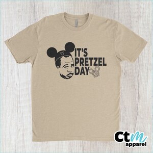 K&ouml;nnte beinhalten: Ein beiges T-Shirt mit einem schwarzen Aufdruck einer Person mit Mickey-Mouse-Ohren und dem Text "It's Pretzel Day" mit einem Brezel.