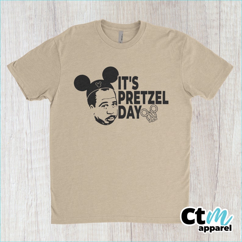 K&ouml;nnte beinhalten: Ein beiges T-Shirt mit einem schwarzen Aufdruck einer Person mit Mickey-Mouse-Ohren und dem Text "It's Pretzel Day" mit einem Brezel.