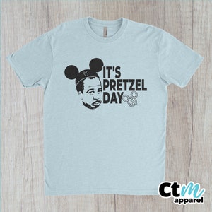 K&ouml;nnte beinhalten: Ein hellblaues T-Shirt mit einem schwarzen Aufdruck einer Person mit Mickey-Mouse-Ohren und dem Text "It's Pretzel Day" mit einem Brezel-Motiv.