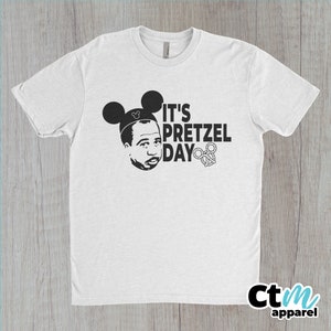 K&ouml;nnte beinhalten: Wei&szlig;es T-Shirt mit einer schwarz-wei&szlig;en Grafik einer Person mit Mickey-Mouse-Ohren und dem Text "It's Pretzel Day".