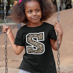 May include: Black t-shirt featuring a leopard print letter "S" with a white border. The child is wearing grey joggers and pink trainers. The child is on a swing.