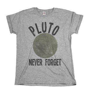 Può includere: Una maglietta grigia con il testo "PLUTO NEVER FORGET" in lettere nere. Un cerchio verde con una texture usurata è sopra il testo.