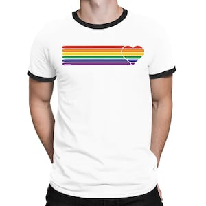 Puede incluir: Camiseta blanca con cuello de canalé negro con un diseño de rayas arcoíris con forma de corazón al final de las rayas.