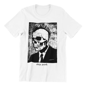 Può includere: T-shirt bianca con una grafica in bianco e nero di uno scheletro con un mohawk che indossa un abito e una cravatta. Il testo "stay punk" è sotto la grafica.