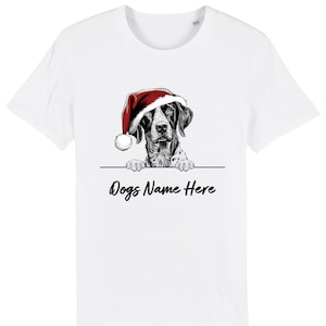 Pode incluir: Camiseta branca com um cachorro preto e branco usando um gorro de Papai Noel. O cachorro está espreitando por cima de uma linha preta com o texto "Dogs Name Here" em uma fonte cursiva. Um design festivo de férias.