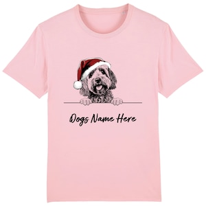 Può includere: T-shirt rosa chiaro con un'illustrazione dettagliata di un cane con un cappello di Babbo Natale. Il testo "Dogs Name Here" è sotto il cane. La maglietta è di colore uniforme e personalizzabile.