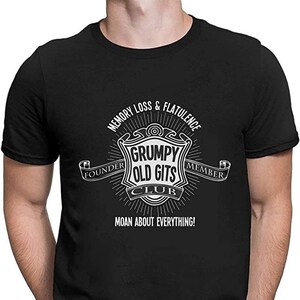 Può includere: Maglietta nera con una grafica bianca di uno stemma che dice "Grumpy Old Gits Club". Il testo "Memory Loss & Flatulence" è sopra lo stemma e "Founder" e "Member" sono su entrambi i lati dello stemma. Il testo "Moan About Everything!" è sotto lo stemma.