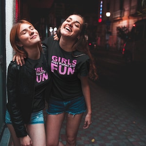 May include: Two friends wearing black t-shirts with pink text. The text on the shirts reads "Girls Just Wanna Have Fun". The friends are smiling and laughing together in a city street at night.