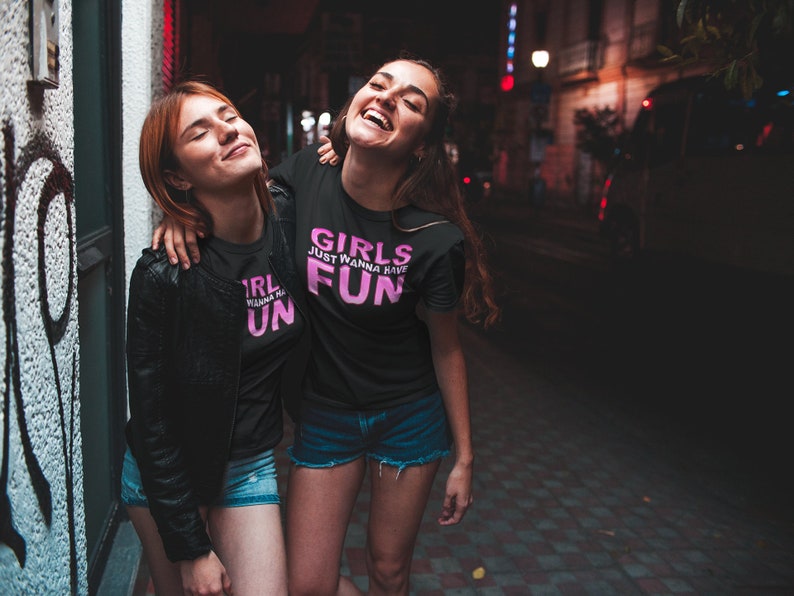 May include: Two friends wearing black t-shirts with pink text. The text on the shirts reads "Girls Just Wanna Have Fun". The friends are smiling and laughing together in a city street at night.