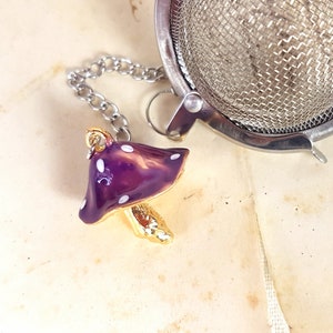 Peut inclure: Infuseur à thé en forme de champignon violet et blanc avec une tige et une chaîne dorées. L'infuseur est suspendu à une passoire à thé en maille argentée.