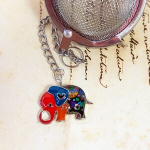 Op de afbeelding: Een thee-ei van roestvrij staal met een decoratieve olifantenbedel. De olifantenbedel is beschilderd met rode, blauwe, oranje en zwarte emaille en heeft een bloemenpatroon. Het thee-ei heeft een ketting en een gaasmandje.
