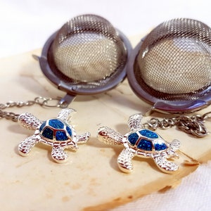 Pode incluir: Dois infusores de chá em aço inoxidável com um pingente de tartaruga. Os infusores têm uma cesta de malha e uma corrente. Os pingentes de tartaruga são prateados com uma concha azul brilhante. Em uma superfície marrom clara.
