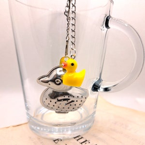 Peut inclure: Une infuseur à thé en argent en forme de canard avec un canard en caoutchouc jaune sur le dessus. L'infuseur est suspendu à une chaîne en argent dans une tasse en verre transparente.