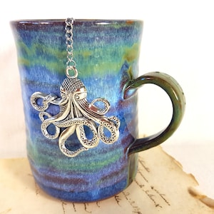 Könnte beinhalten: Eine blaue und grüne Keramiktasse mit einem silbernen Tintenfisch-Tee-Infuser, der am Rand hängt.