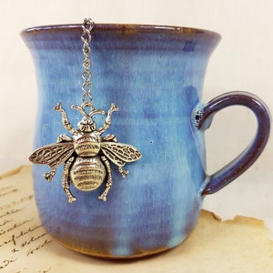 Pode incluir: Uma caneca de cerâmica azul com uma borda e uma alça marrons. Um infusor de chá de abelha prateado pendura na borda da caneca.
