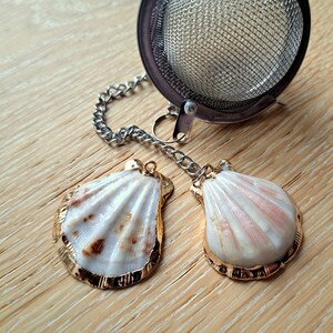 Op de afbeelding: Twee witte schelpen met gouden randen bevestigd aan een zilveren ketting en een zilveren thee-infuser. De schelpen liggen op een houten oppervlak.
