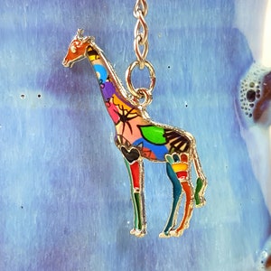Könnte beinhalten: Ein bunter Giraffen-Anhänger mit einer silbernen Kette. Die Giraffe ist aus Metall und hat eine mehrfarbige Emaille-Oberfläche. Der Anhänger ist perfekt, um einem Schlüsselanhänger, einer Handtasche oder einem Rucksack einen Hauch von Verspieltheit zu verleihen.