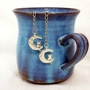 Puede incluir: Una taza de cerámica azul con borde y asa marrón, que contiene dos infusores de té plateados con forma de gato y luna. Los infusores cuelgan de cadenas y presentan una luna creciente con una silueta de gato.