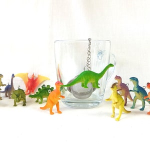 Könnte beinhalten: Ein Set bunter Dinosaurier-Tee-Infuser, darunter ein grüner Stegosaurus, ein roter Pterodactylus, ein blauer Triceratops, ein gelber Brachiosaurus, ein lila Velociraptor, ein grüner Ankylosaurus, ein orangefarbener Tyrannosaurus Rex, ein grüner Parasaurolophus, ein blauer Spinosaurus und ein rot-orangefarbener Carnotaurus. Die Dinosaurier hängen an einem Metall-Tee-Infuser.