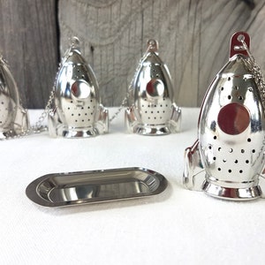 Könnte beinhalten: Vier silberne, raketenförmige Tee-Infuser mit Ketten und einem silbernen Tablett. Die Infuser haben ein rundes Loch vorne und sind mit kleinen Löchern perforiert.