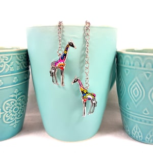 Könnte beinhalten: Zwei Giraffenanhänger aus silberfarbenem Metall, die an silbernen Ketten hängen. Jede Giraffe ist mit farbigen Emaille-Details in Rosa, Blau, Gelb und Schwarz verziert und trägt ein kleines rotes Herz auf dem Körper der Tierfigur.