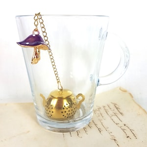 Op de afbeelding: Een gouden theepotvormige thee-infuser met een paarse paddenstoelbedel die aan een gouden ketting hangt. De infuser bevindt zich in een heldere glazen mok met een handvat.