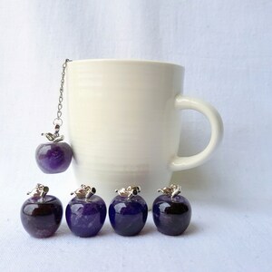Può includere: Cinque infusori per tè a forma di mela in ametista, uno appeso a una catena d'argento su una tazza bianca. Le mele di ametista sono di un colore viola scuro.
