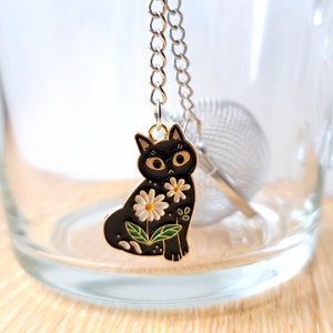 Può includere: Un infusore da tè a forma di gatto nero con una catena dorata e fiori bianchi. L'infusore è appeso a una tazza da tè di vetro.