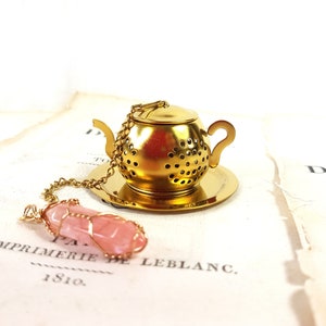 Op de afbeelding: Gouden theepotvormige thee-infuser met een roze kristallen hanger aan een ketting. De infuser heeft een deksel en een schoteltje. De kristallen hanger is omwikkeld met goudkleurig draad.