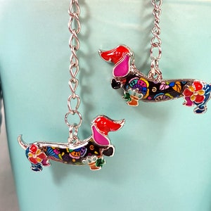 Könnte beinhalten: Zwei silberne Anhänger-Ohrringe mit einem Dackel-Design. Der Dackel ist schwarz mit bunten Blumenmustern und rosa Ohren. Die Ohrringe hängen an silbernen Ketten.